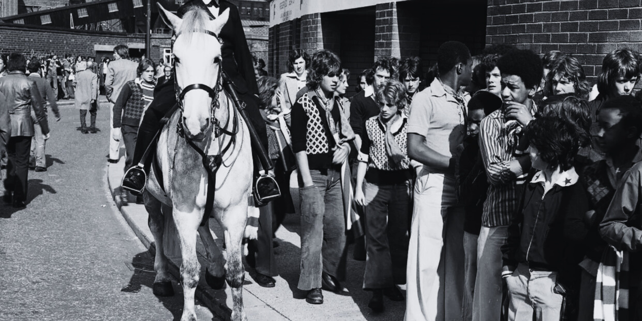 Old Trafford, 1975: Manchester United fans in flares, big collars, scarves, boots and sharp ’70s cuts queue for the Stretford End - admission 65p - as mounted police keep order.