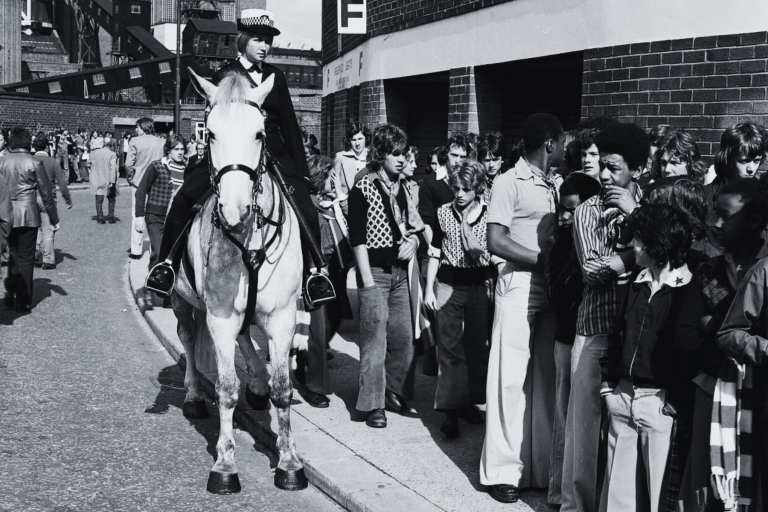 Old Trafford, 1975: Manchester United fans in flares, big collars, scarves, boots and sharp ’70s cuts queue for the Stretford End - admission 65p - as mounted police keep order.